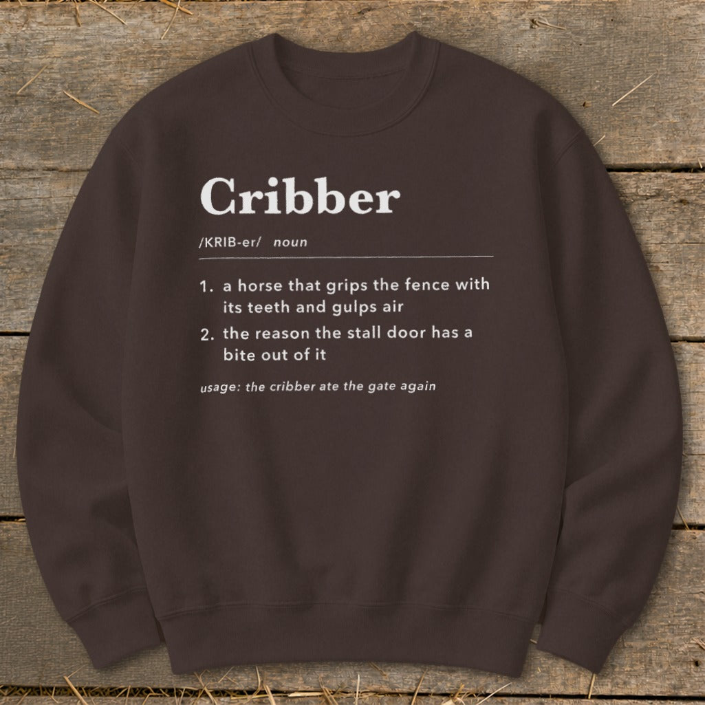 A dark brown crewneck sweatshirt featuring white dictionary-style text defining the word cribber laid on a wooden surface.