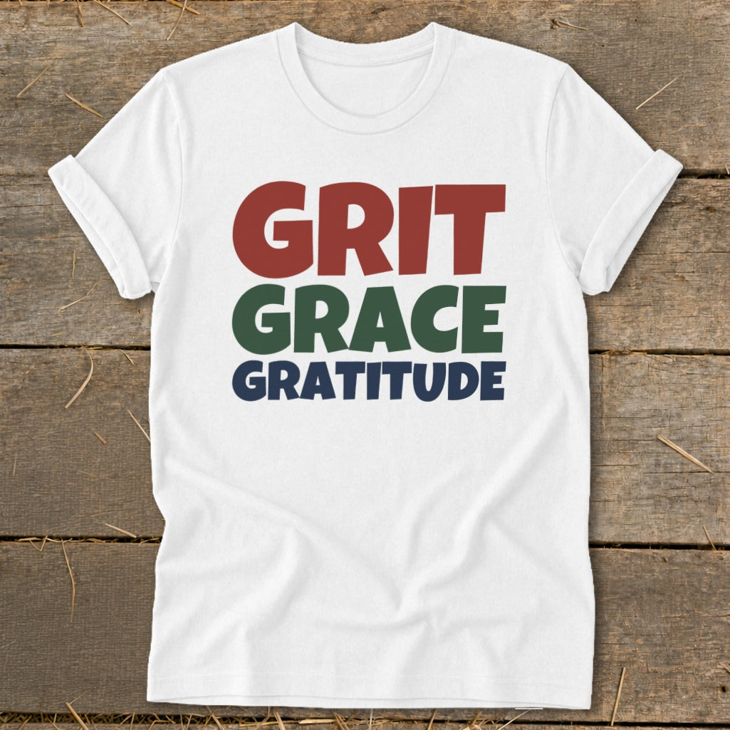 White short-sleeve t-shirt with rolled cuffs featuring stacked text reading GRIT, GRACE, GRATITUDE in red, green, and navy.