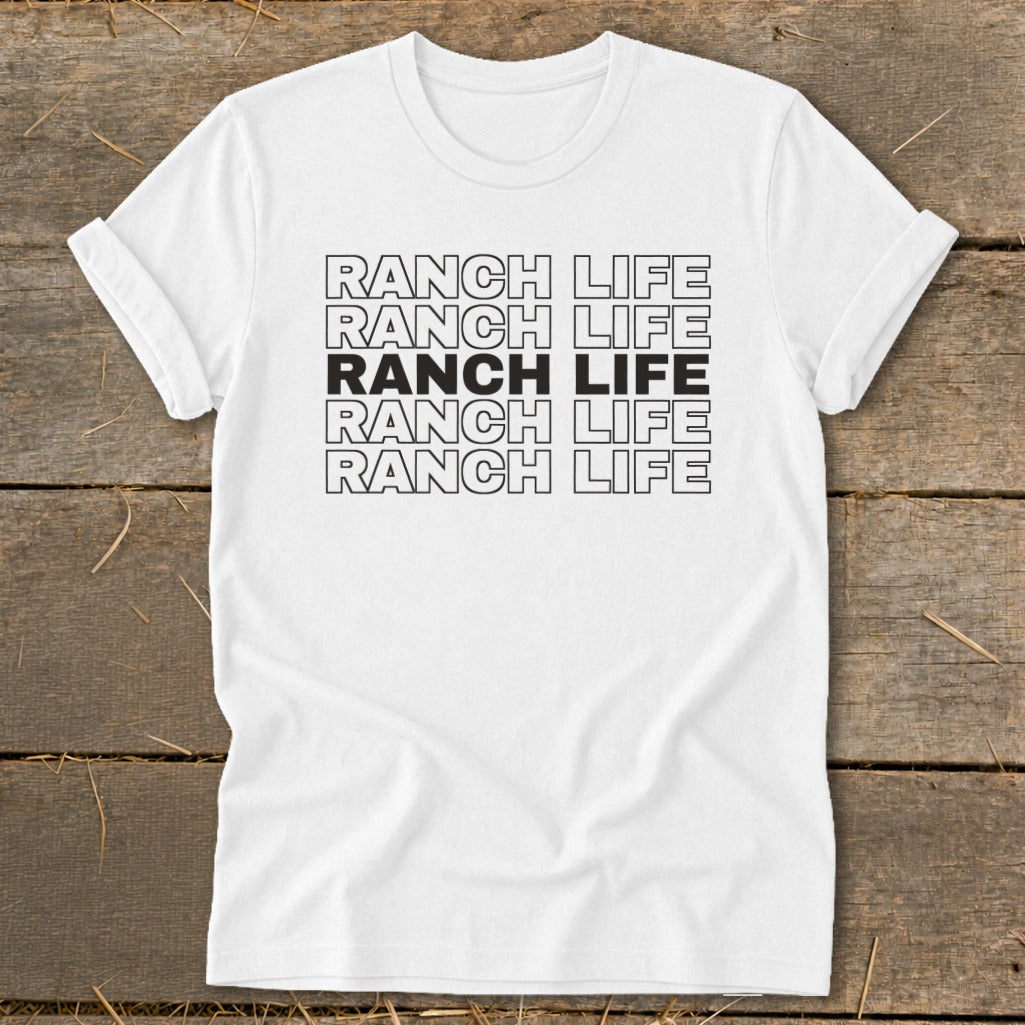 A white crewneck t-shirt with rolled sleeves lies flat on a rustic wooden surface, featuring centered black text reading RANCH LIFE repeated in five stacked rows, with the middle row solid black.