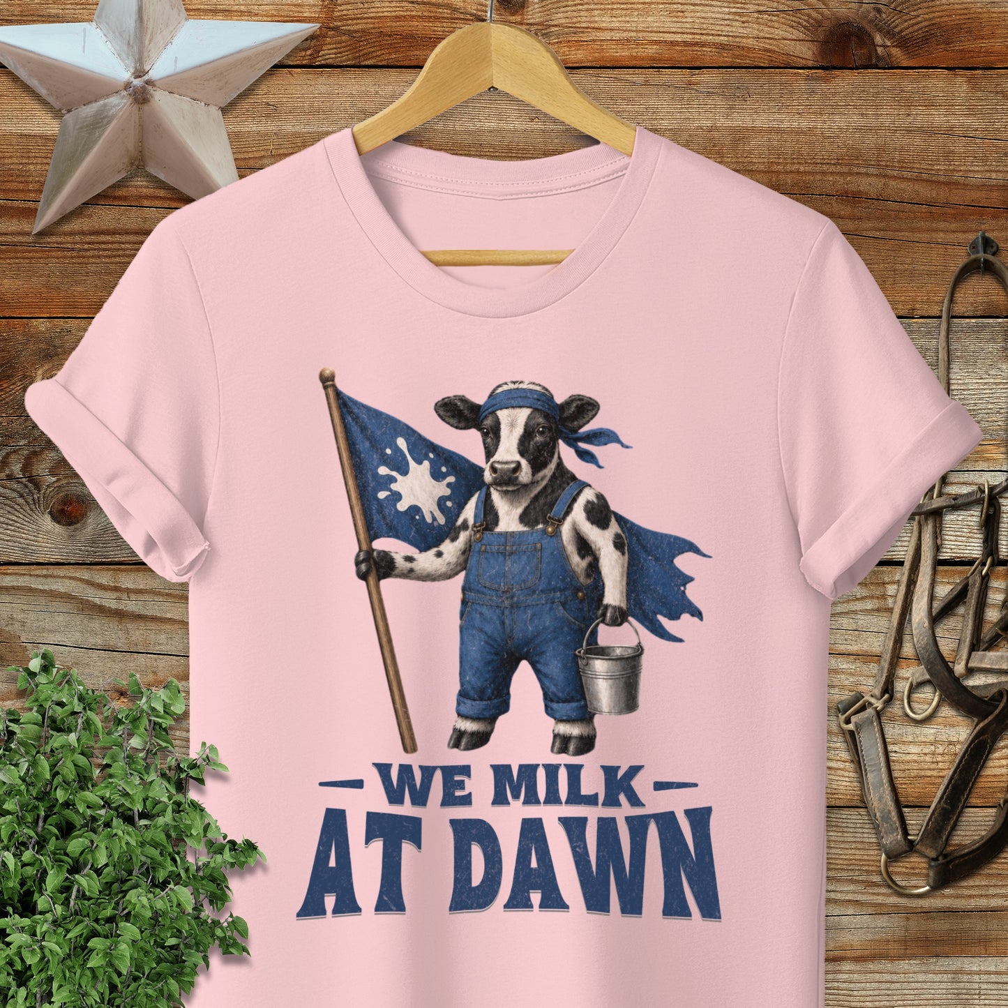 Cow wearing overalls and cape, holding milk pail and flag with "We Milk at Dawn" text.