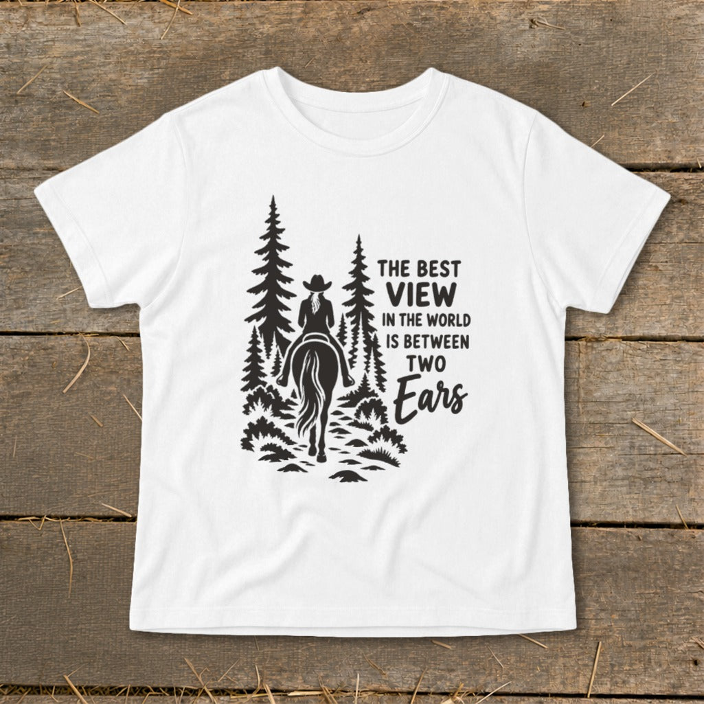 A white t-shirt featuring a black graphic of a rider on a horse surrounded by pine trees, with the text "The best view in the world is between two ears."