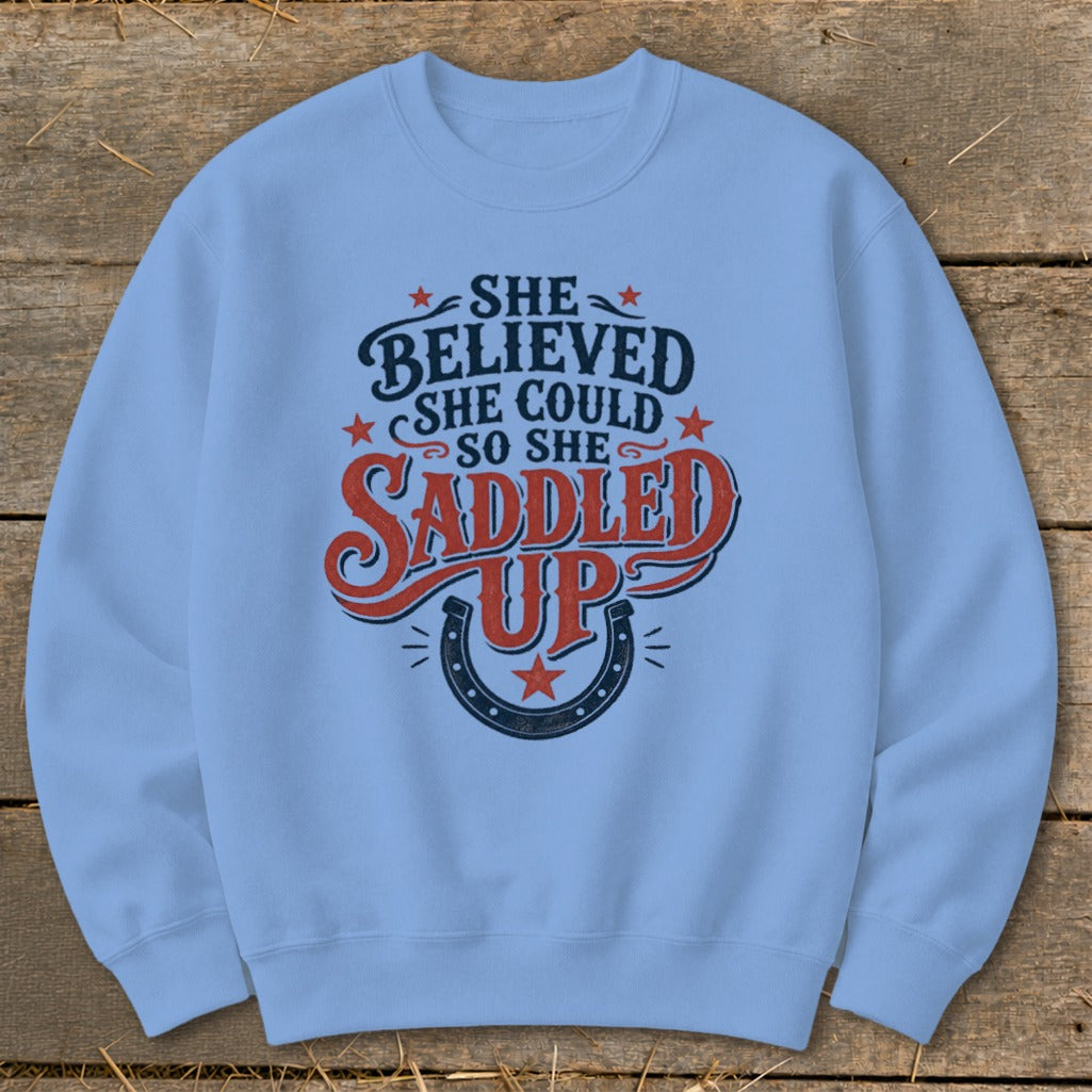 Light blue sweatshirt with "She believed she could so she saddled up" graphic and horseshoe.