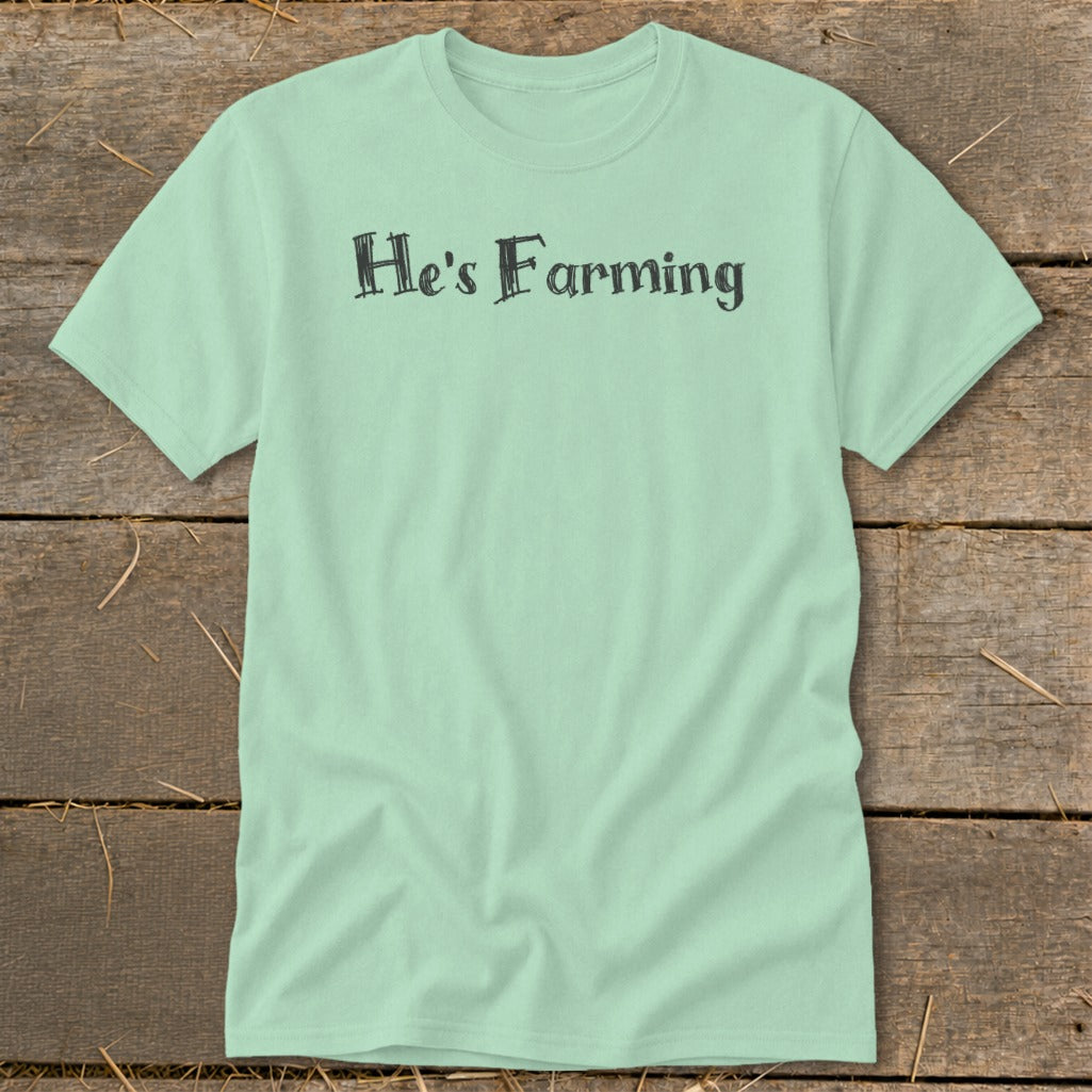 Light green t-shirt with "He's Farming" text in dark grey, on a wooden background.