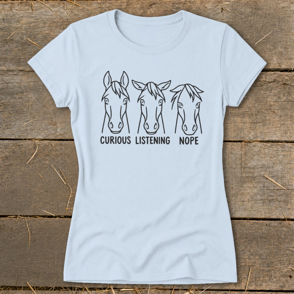 A light blue crewneck t-shirt featuring three horse head illustrations with the text "Curious, Listening, Nope" below them, laid flat on a rustic wooden background.