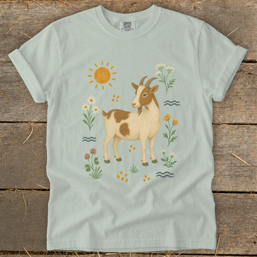 Light blue t-shirt with a brown and white goat surrounded by flowers, sun, and waves.