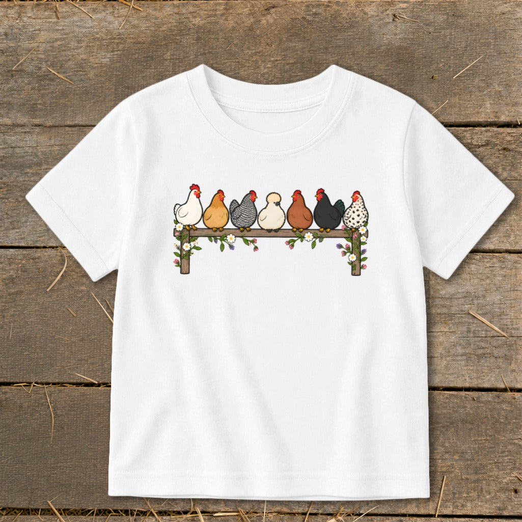 A white children’s t-shirt displays an illustration of seven colorful chickens lined up on a wooden fence decorated with flowers.