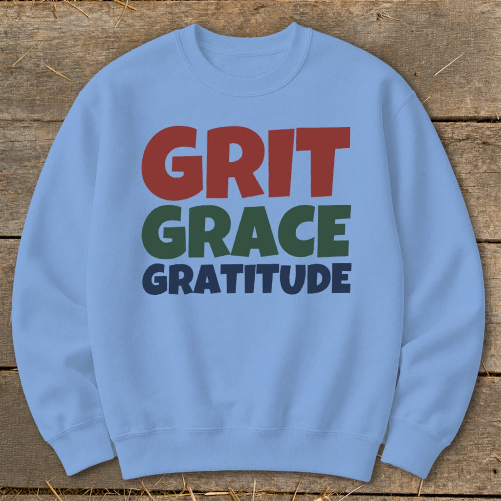 Light blue crewneck sweatshirt with colorful text reading GRIT GRACE GRATITUDE laid on wood.