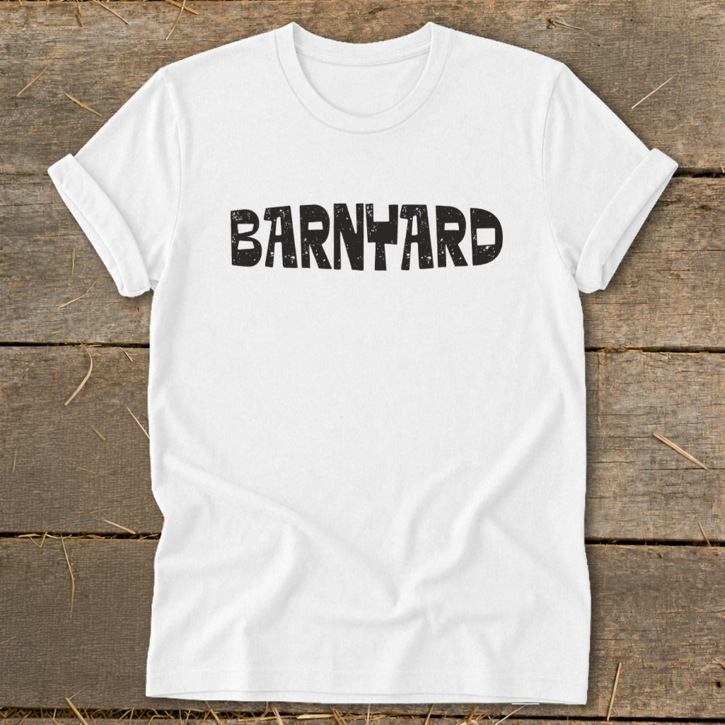 A white short-sleeved t-shirt with rolled cuffs rests flat on a rustic wooden surface, featuring the distressed black text "BARNYARD" centered on the chest.