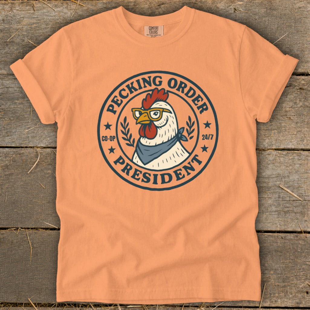Peach t-shirt with a circular graphic of a chicken wearing glasses and a bandana, "Pecking Order President 24/7" text.