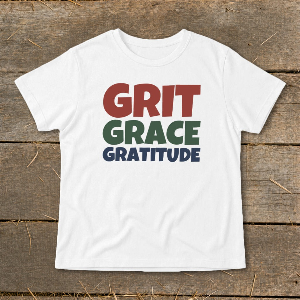A white short-sleeve t-shirt with multicolored text reading GRIT GRACE GRATITUDE centered on a wooden background.