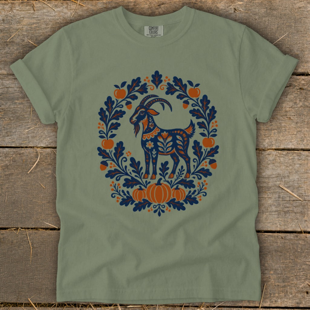 A sage green t-shirt featuring a folk-art goat illustration surrounded by autumn leaves, apples, and pumpkins.