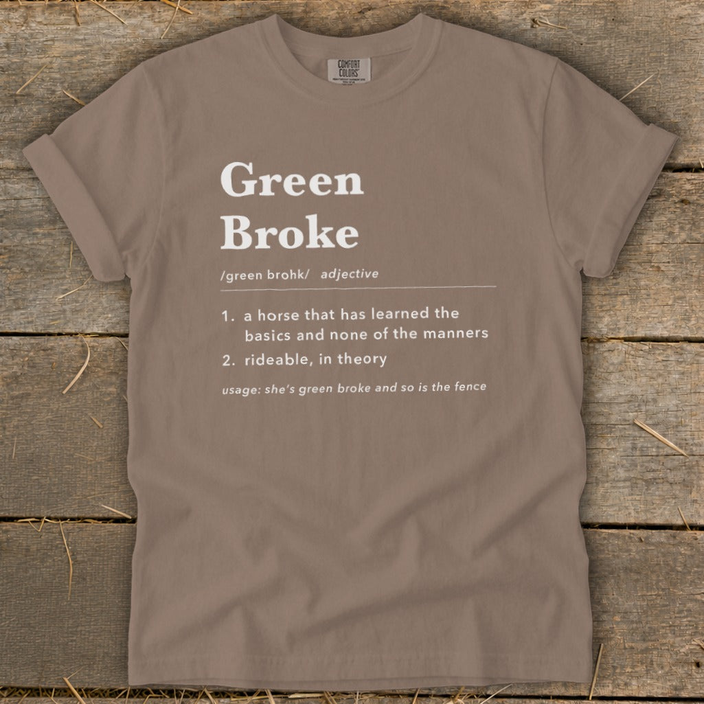 A brown t-shirt featuring white text that defines the adjective Green Broke on a rustic wooden background.