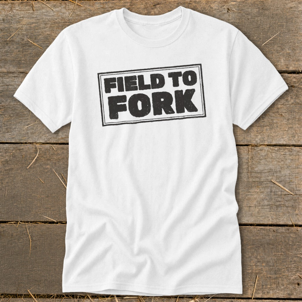 A white t-shirt with black text that reads "FIELD TO FORK" inside a rectangular border, laid flat on a wooden surface.