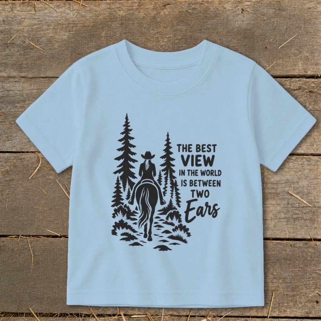 Light blue t-shirt featuring a silhouette of a person on a horse riding through trees with text reading, The best view in the world is between two ears.