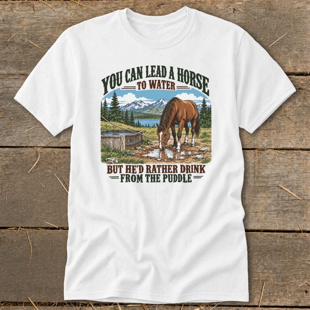 A white t-shirt featuring a graphic of a horse drinking from a puddle with mountains and the text "You can lead a horse to water, but he'd rather drink from the puddle."
