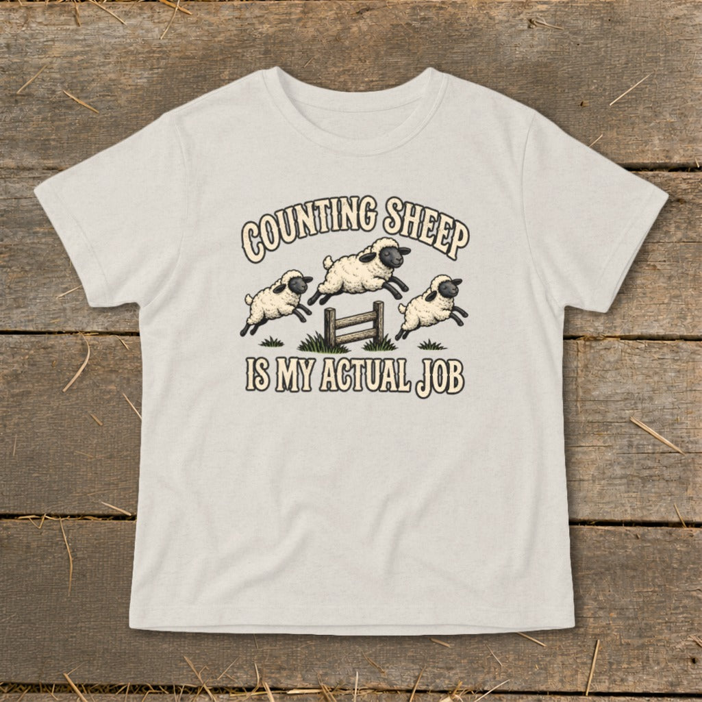 Light beige t-shirt with cartoon sheep jumping a fence, reading "Counting Sheep Is My Actual Job".