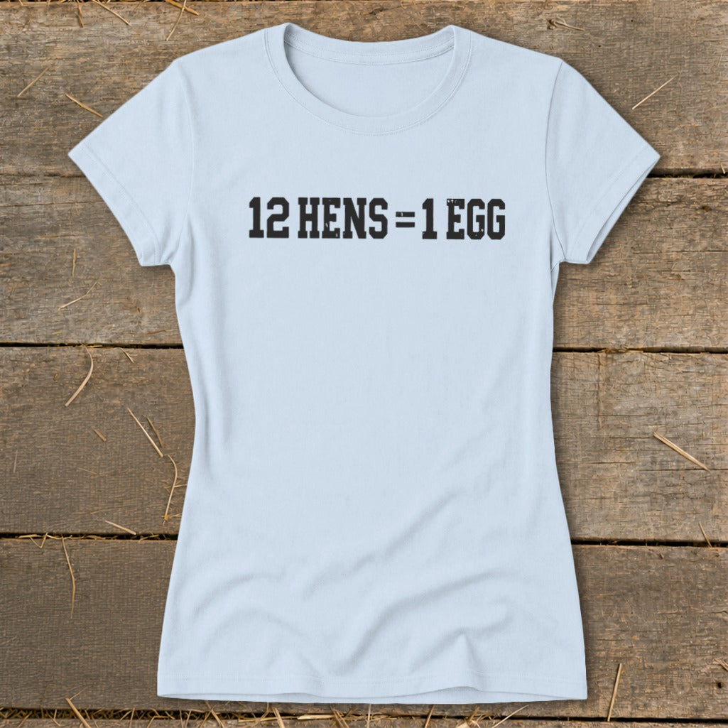 Light blue women's short-sleeve t-shirt with the black text "12 HENS = 1 EGG" centered on the chest, laid flat on a rustic wooden background.