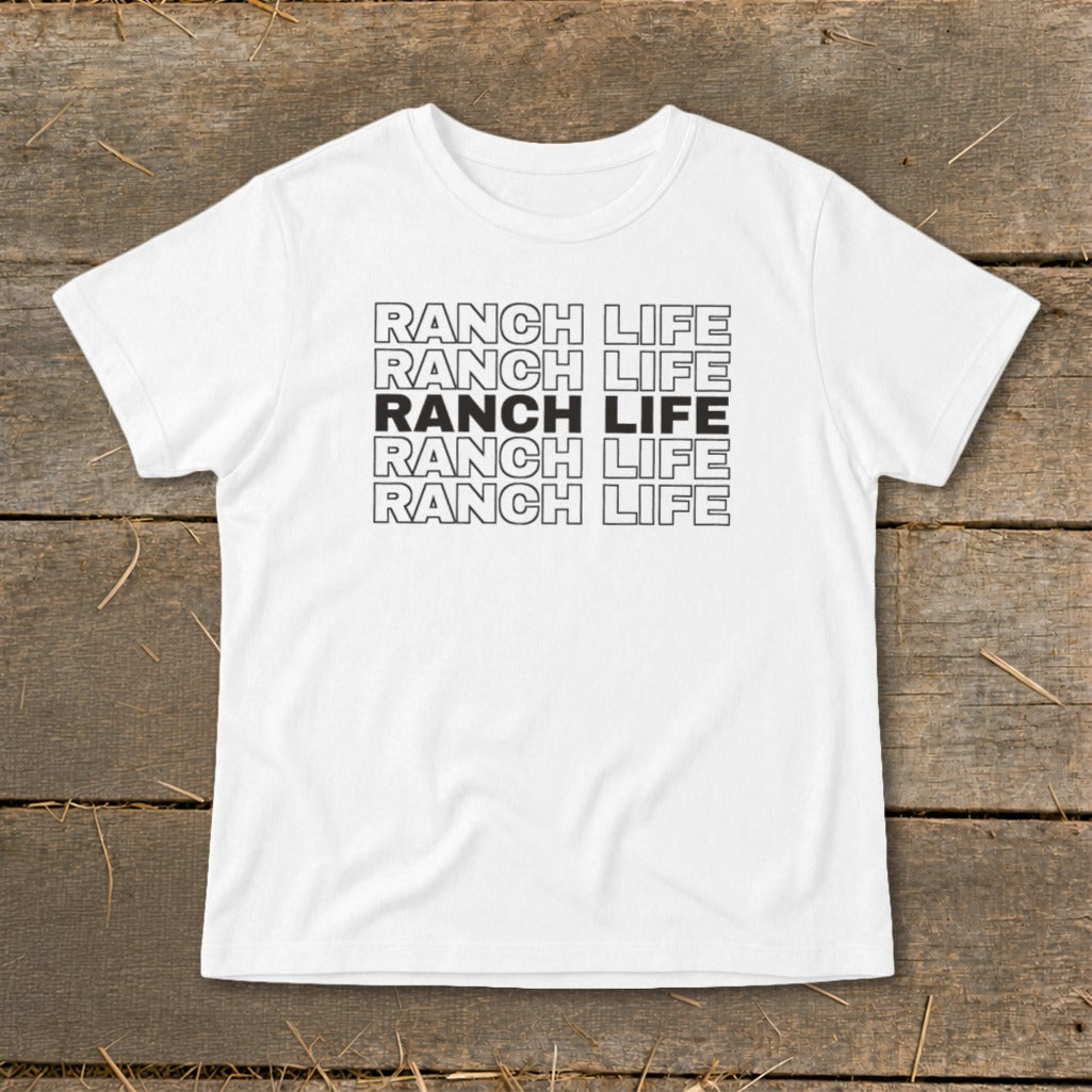 A white short-sleeve t-shirt with five stacked lines of text reading RANCH LIFE on a wooden surface.