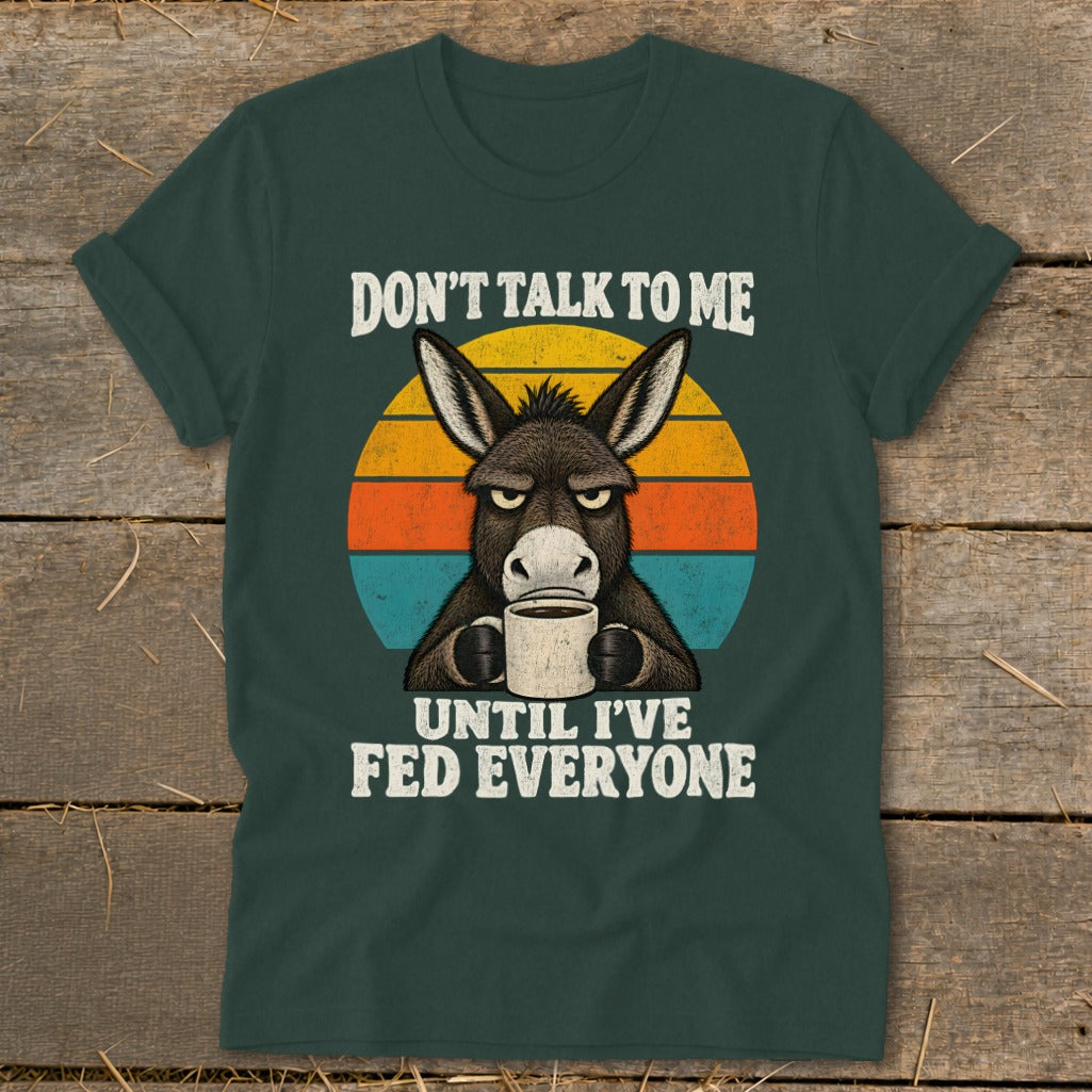 A dark green t-shirt with a graphic of a grumpy donkey holding a coffee mug and the text Don't Talk To Me Until I've Fed Everyone against a vintage sunset background.