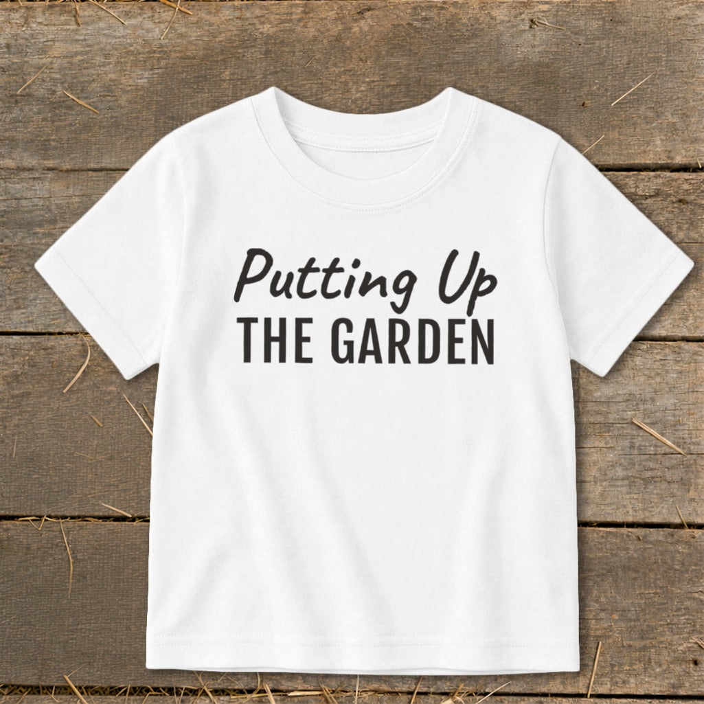 White t-shirt with black text "Putting Up THE GARDEN" on a wooden background.