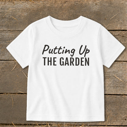 White t-shirt with black text "Putting Up THE GARDEN" on a wooden background.