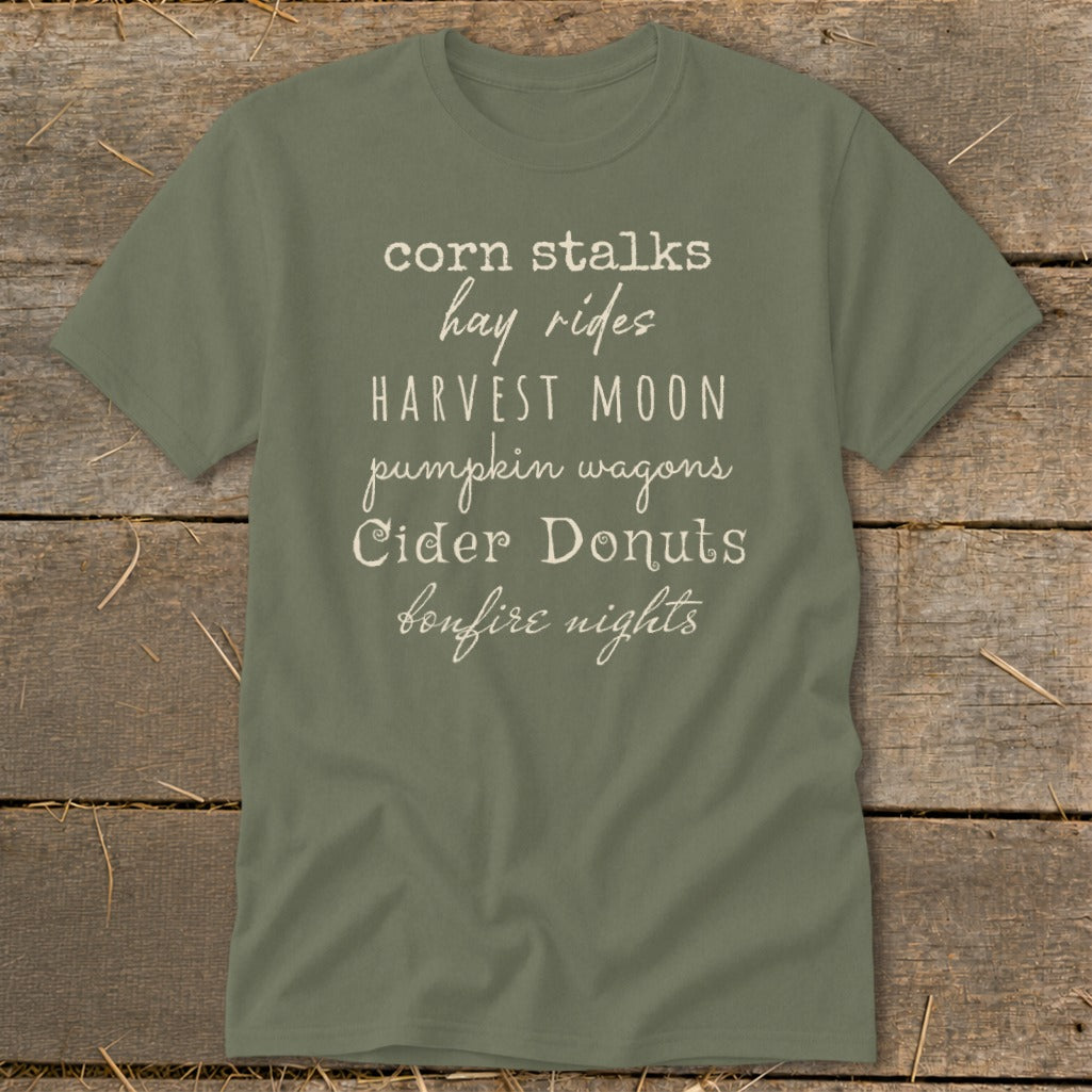 Green t-shirt with white text listing autumn activities: corn stalks, hay rides, harvest moon, pumpkin wagons, cider donuts, bonfire nights.