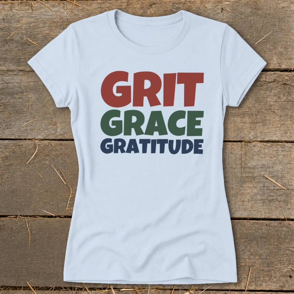 Light blue t-shirt featuring the stacked text GRIT GRACE GRATITUDE displayed on a wooden background.