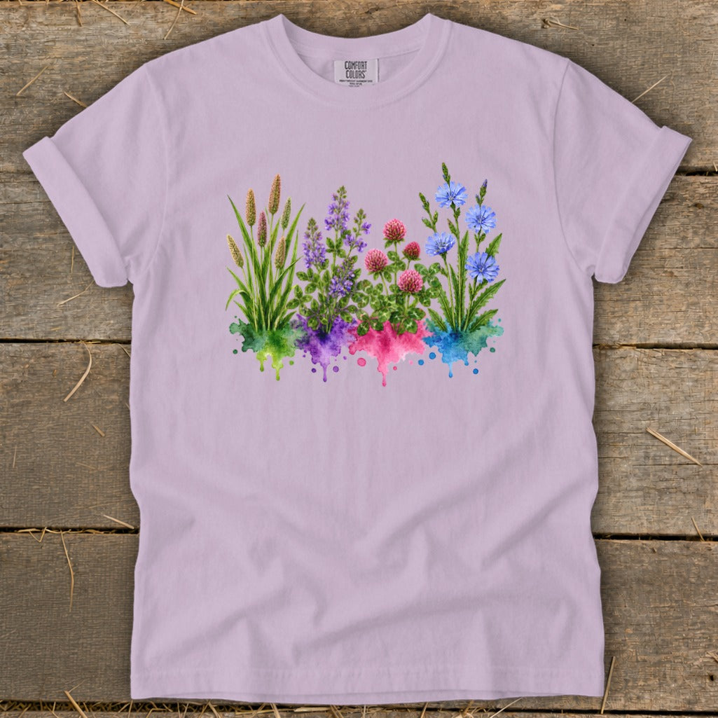 A light purple short-sleeve t-shirt lies flat on a rustic wooden surface, featuring a colorful watercolor wildflower graphic.