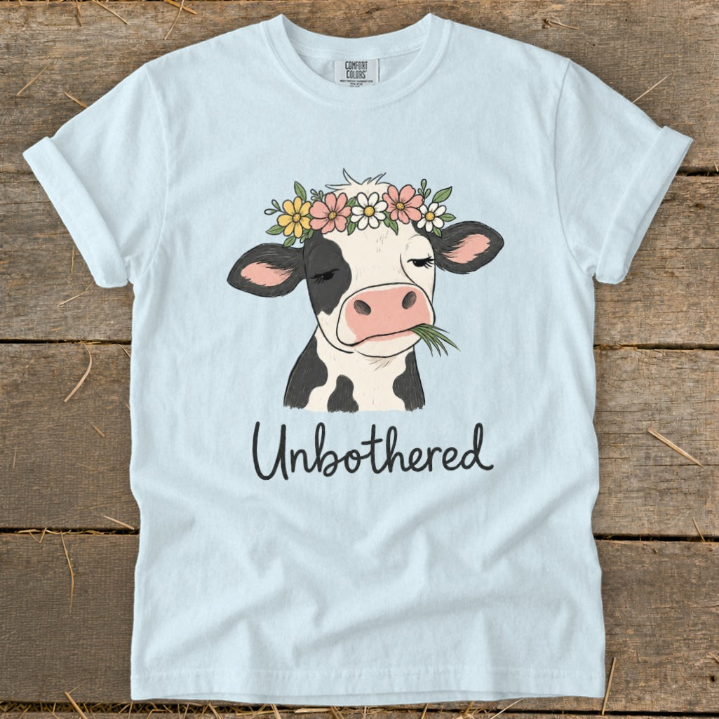 Light blue t-shirt with a cow wearing a flower crown and chewing grass, reads "Unbothered".