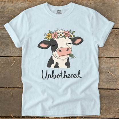 Light blue t-shirt with a cow wearing a flower crown and chewing grass, reads "Unbothered".