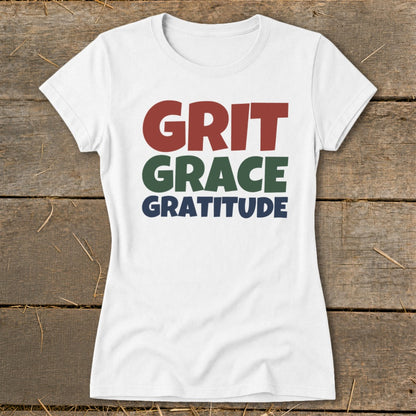 White short-sleeved t-shirt with multicolored stacked text reading GRIT GRACE GRATITUDE laid flat on wood.