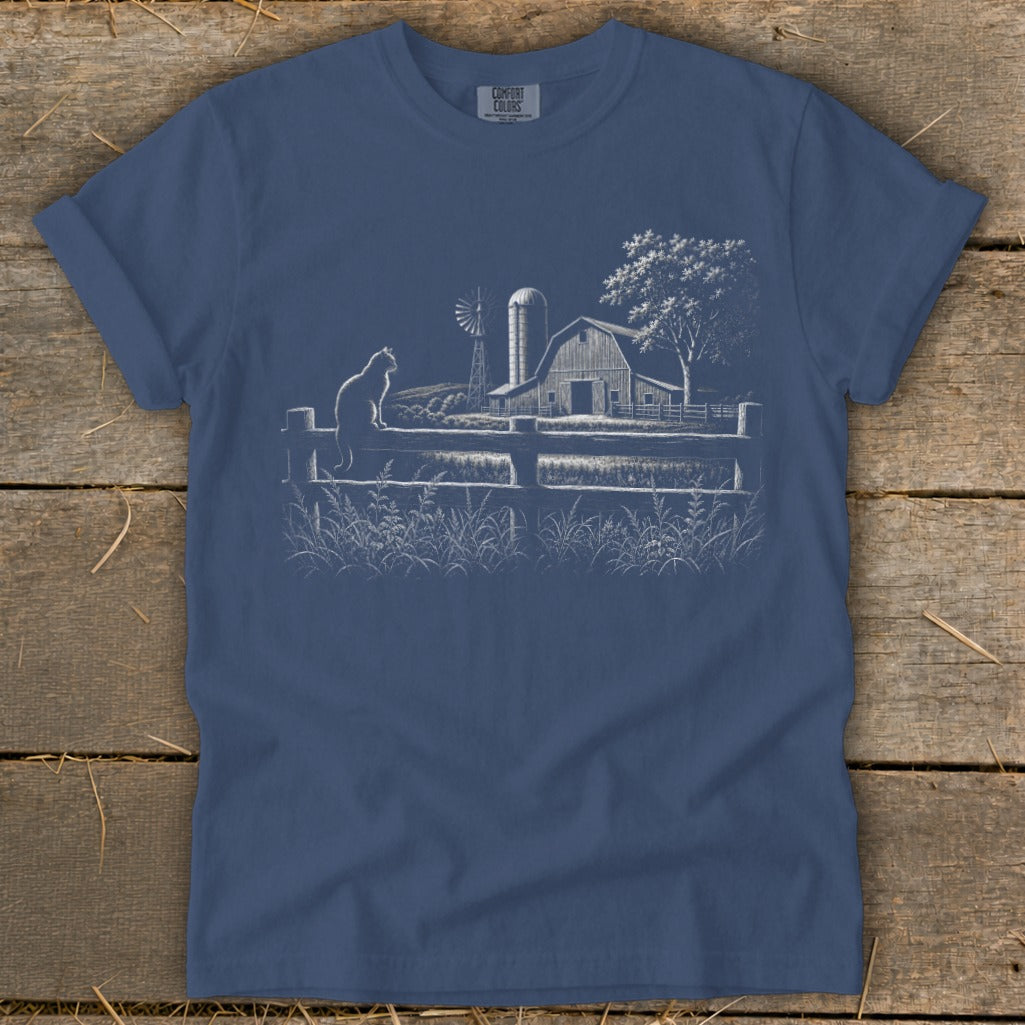 White line drawing on blue t-shirt of a cat on a fence overlooking a farm scene.