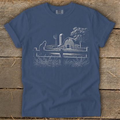 White line drawing on blue t-shirt of a cat on a fence overlooking a farm scene.