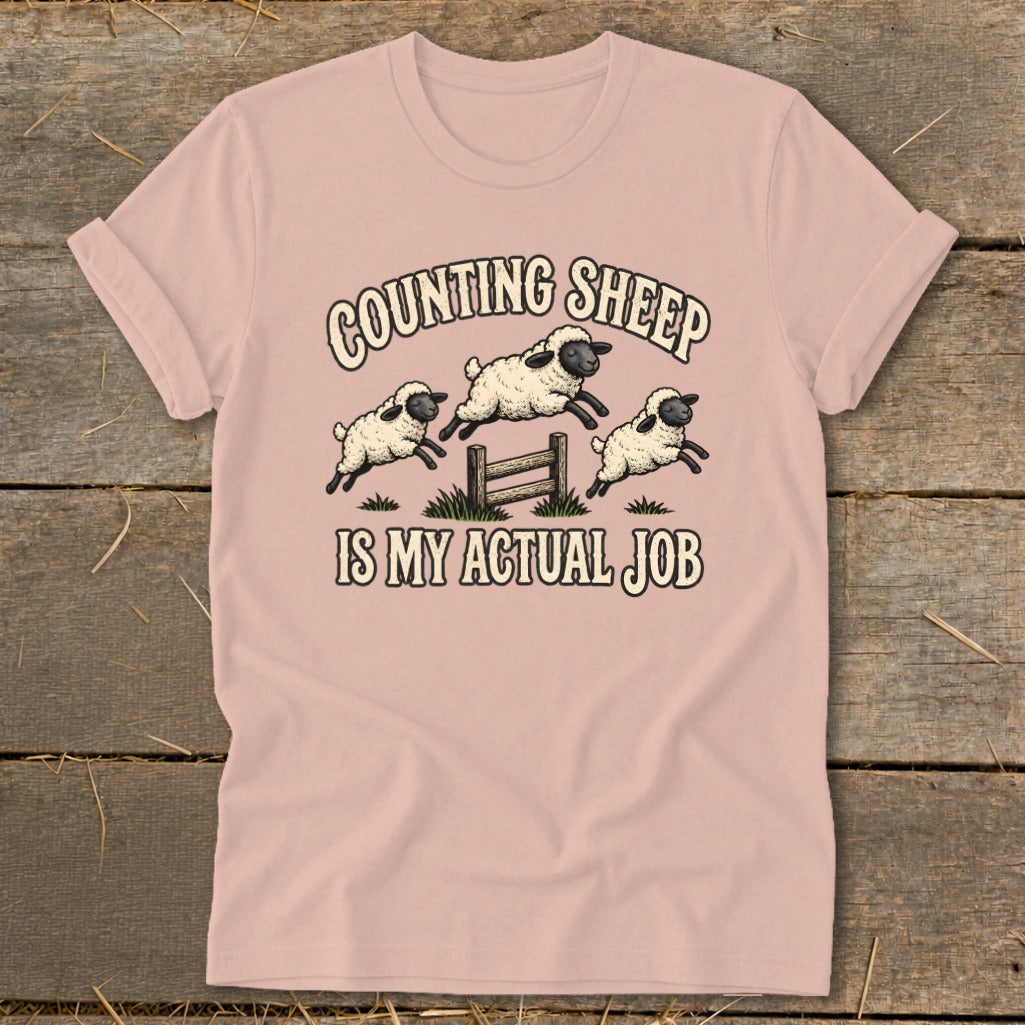 Pink t-shirt with cartoon sheep jumping over a fence, text reads "Counting Sheep Is My Actual Job".
