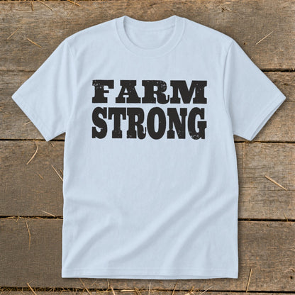Light blue short-sleeve t-shirt with black distressed text reading "FARM STRONG" laid flat on weathered wood planks.