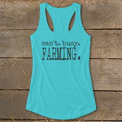Turquoise tank top with "can't. busy. FARMING." text on wooden background.