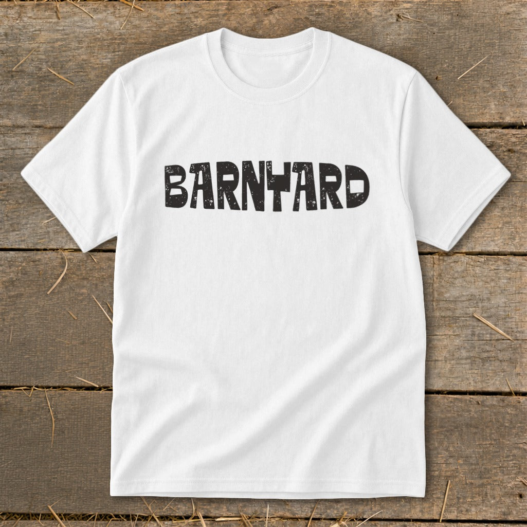 A white crewneck t-shirt lies flat on a rustic wooden surface, featuring the black distressed text BARNYARD centered on the chest.