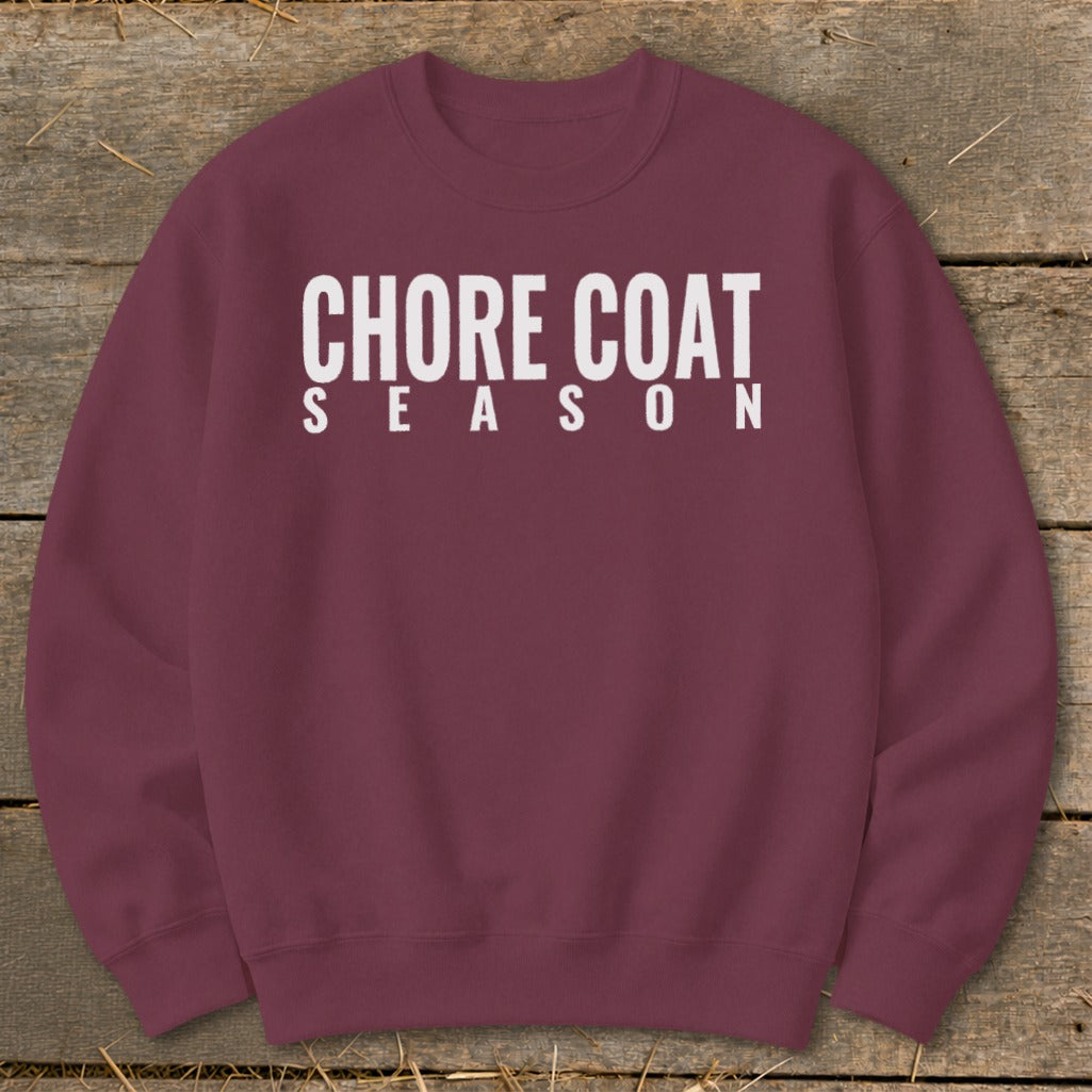 Maroon sweatshirt with white text reading "CHORE COAT SEASON" on a wooden background.