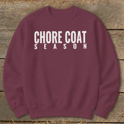 Maroon sweatshirt with white text reading "CHORE COAT SEASON" on a wooden background.