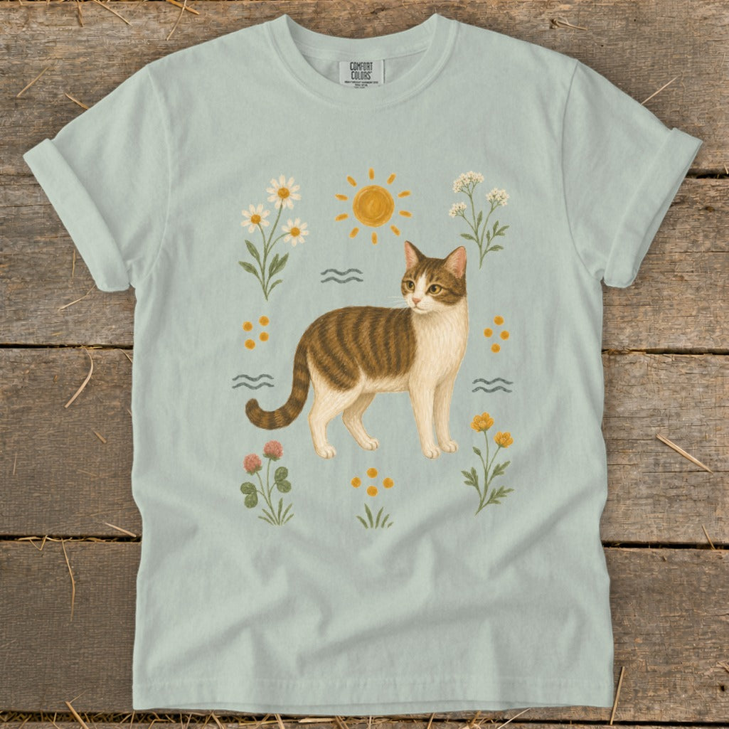 Light blue t-shirt with a brown and white tabby cat surrounded by sun, wildflowers, and clover.