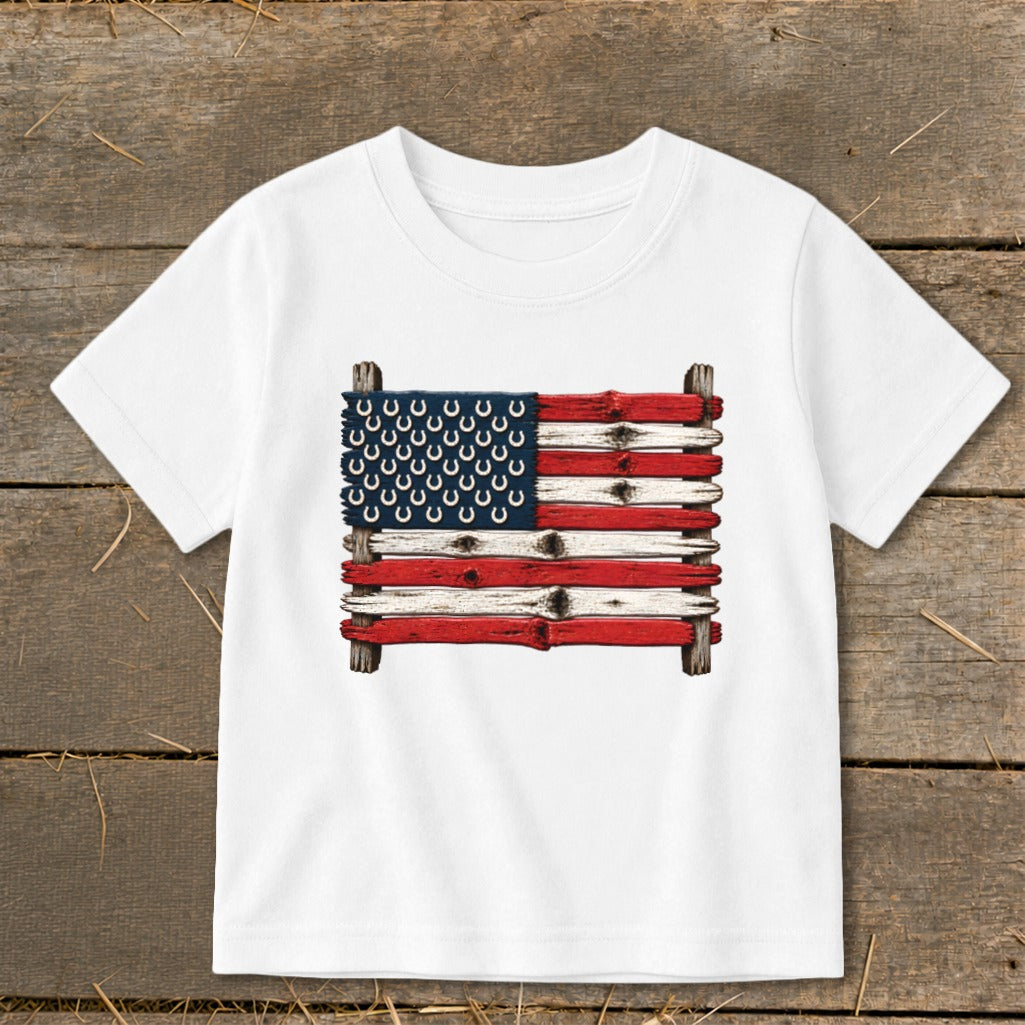 A white short-sleeved children's t-shirt features a graphic of the American flag made of painted wooden planks and horseshoes.
