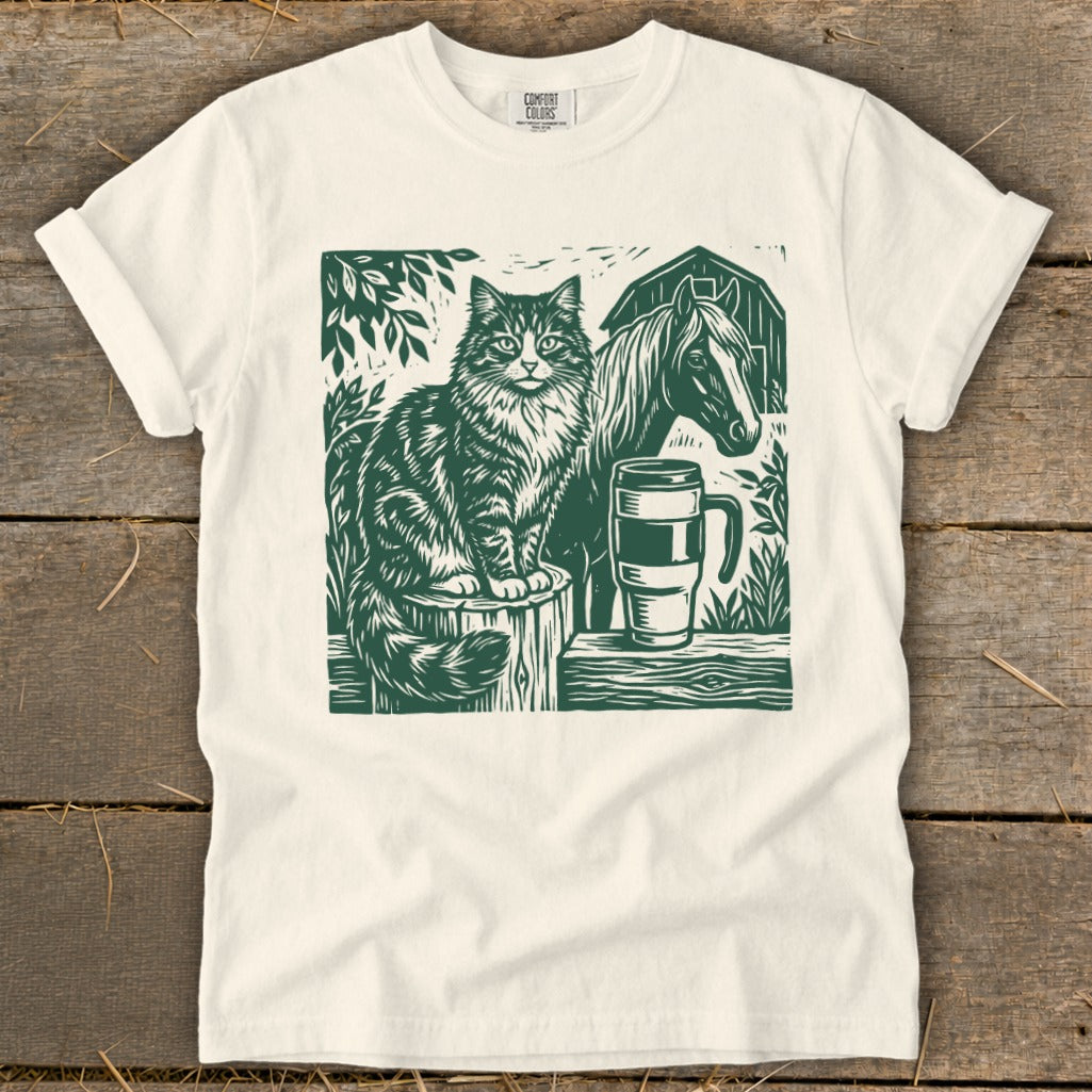 Green linocut design of a cat, horse, mug, and barn on a cream t-shirt.