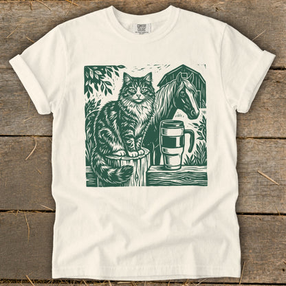 Green linocut design of a cat, horse, mug, and barn on a cream t-shirt.