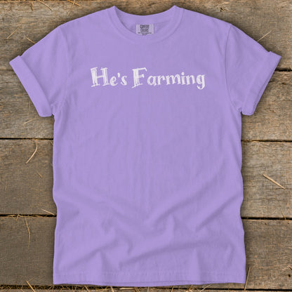 Purple t-shirt with "He's Farming" text on a wooden background.