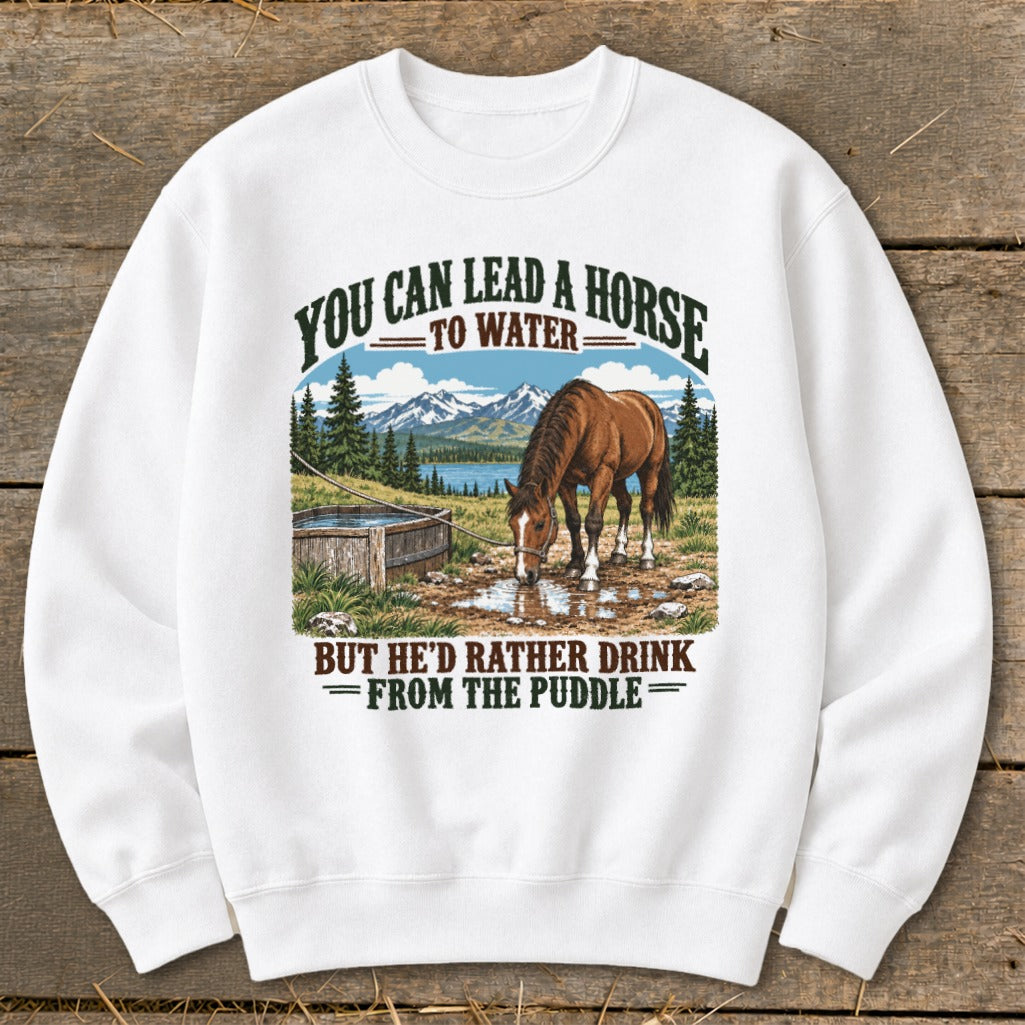 A white crewneck sweatshirt featuring a graphic of a horse drinking from a puddle with mountains and the text You Can Lead a Horse to Water But He'd Rather Drink From the Puddle.