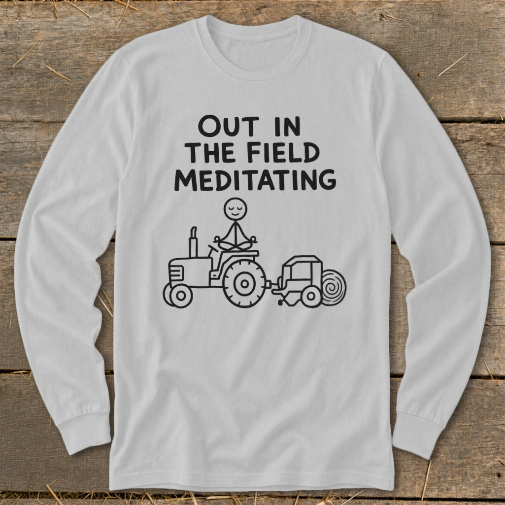 White long-sleeve shirt with text "OUT IN THE FIELD MEDITATING" and a stick figure meditating on a tractor.
