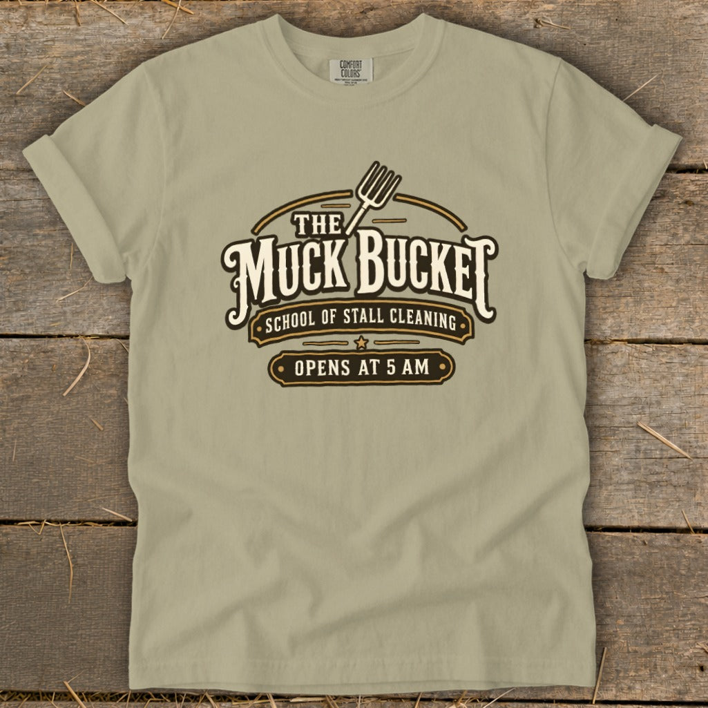 Tan t-shirt with "The Muck Bucket School of Stall Cleaning Opens at 5 AM" logo.