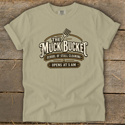 Tan t-shirt with "The Muck Bucket School of Stall Cleaning Opens at 5 AM" logo.