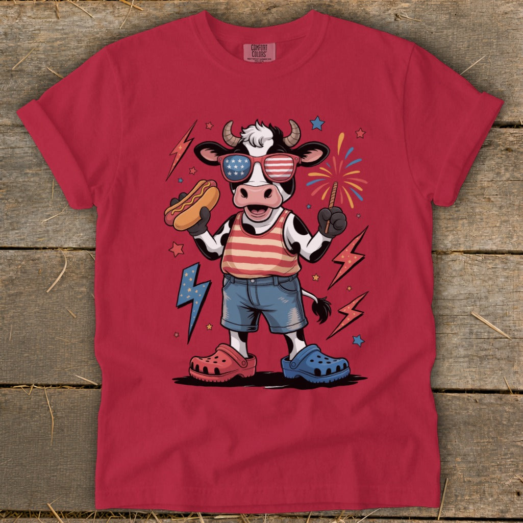Cartoon cow in patriotic attire holding hot dog and sparkler with fireworks.