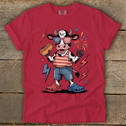 Cartoon cow in patriotic attire holding hot dog and sparkler with fireworks.